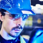 Marmon Keystone A person wearing a blue hard hat, safety glasses, and a reflective vest examines a tablet in an industrial setting, possibly managing tube and pipe supplychain operations, with blurred lights in the background.