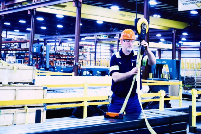 Marmon Keystone A worker in a hard hat and safety gear operates a hoist to lift a heavy metal bar inside an industrial warehouse, highlighting the vital role of metals in the supplychain with shelves, equipment, and yellow railings all around.