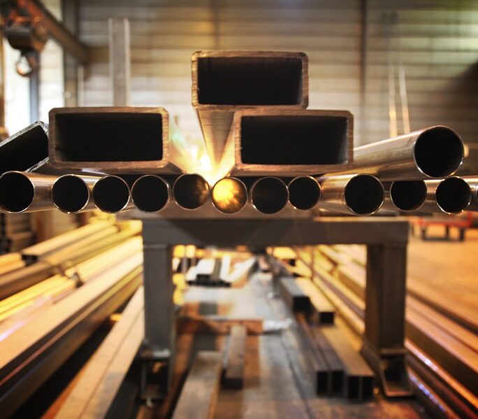 Marmon Keystone A stack of metal pipes and tube bars is arranged in a row inside an industrial warehouse, with the open ends facing the camera and warm lighting illuminating the space—showcasing a reliable metals distributor in the supply chain.
